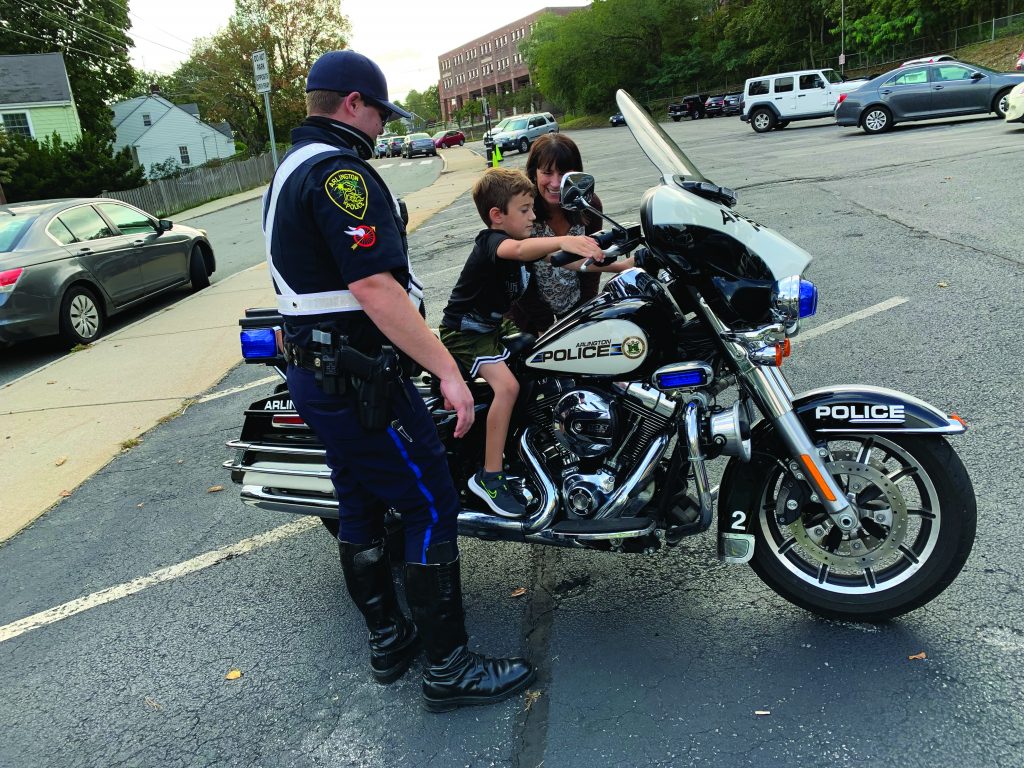 A police officer from Arlington, MA, interacting with children during a "Touch-a-Truck" event. The officer is standing next to a police vehicle, engaging with the community and showcasing the vehicle's features.