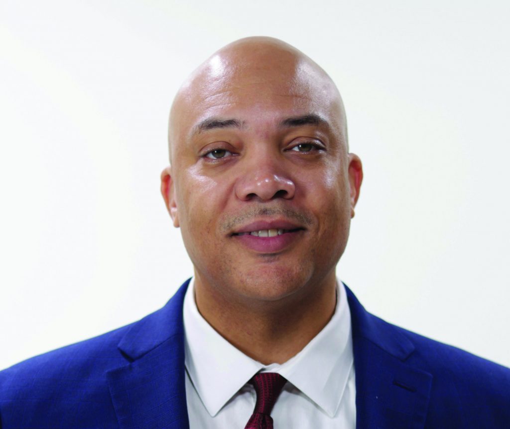Individual in blue suit jacket, white shirt, and red tie posed against plain white background; shaved head, facing forward, expression neutral.
