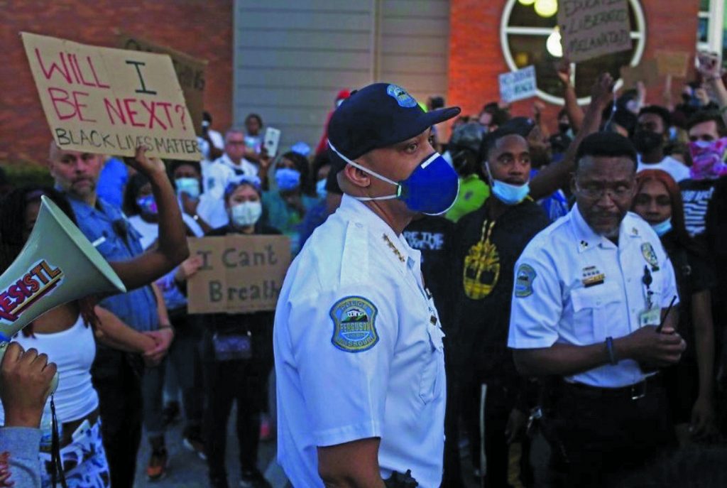 Two uniformed officers stand in crowd of protesters holding signs like 'Will I Be Next?' and 'I Can't Breathe'; one protester speaks through megaphone
