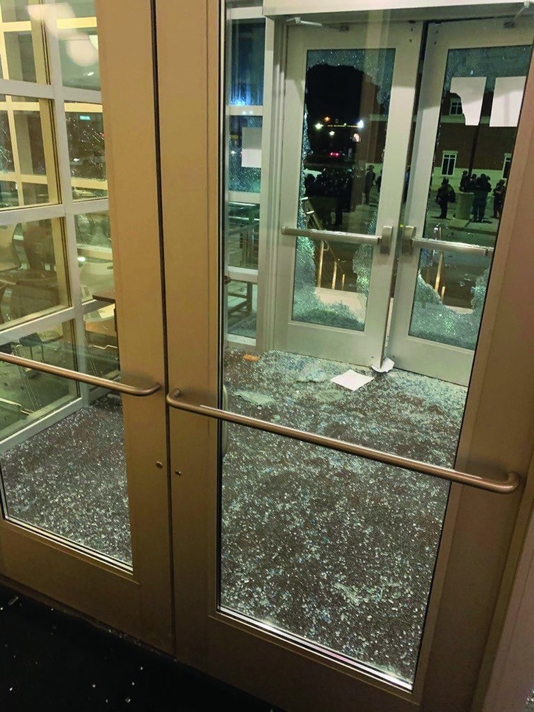 Shattered glass doors with broken windows, scattered shards on floor, paper near entrance; nighttime scene with reflections visible outside