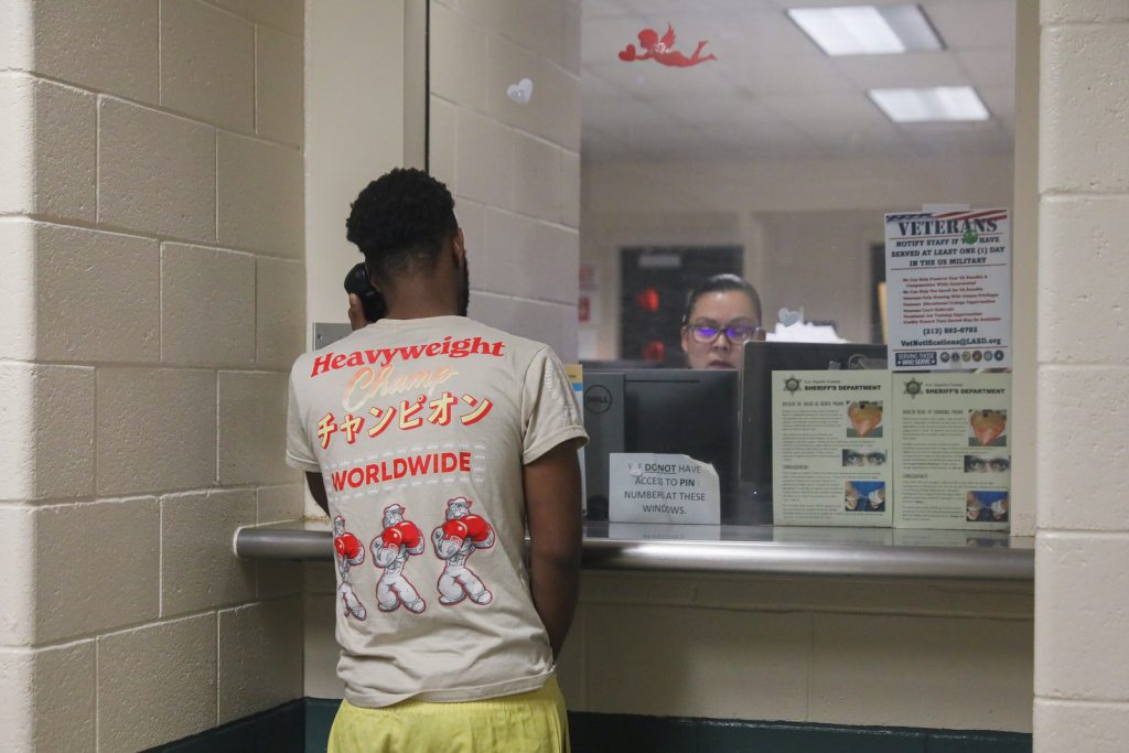 Person in graphic T‑shirt and yellow shorts uses a phone at a jail intake window lined with posted notices, forms, and signage in a tiled hallway area
