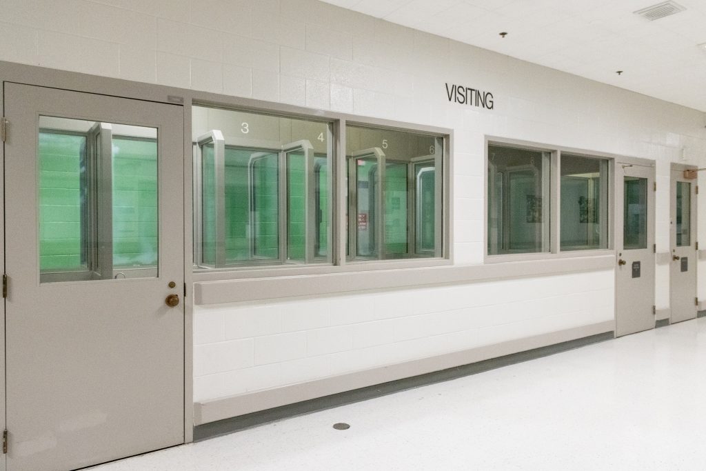 Clean hallway with labeled visiting booths numbered 3 to 6, each with glass partitions and gray framing, alongside secured doors and white tile flooring