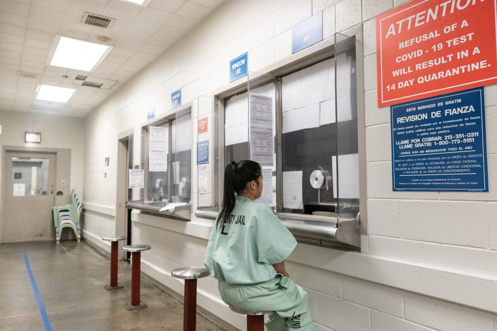 Person in light‑green jail uniform sits on a stool at a row of service windows lined with forms and signs, including large COVID‑19 and bail‑information notices