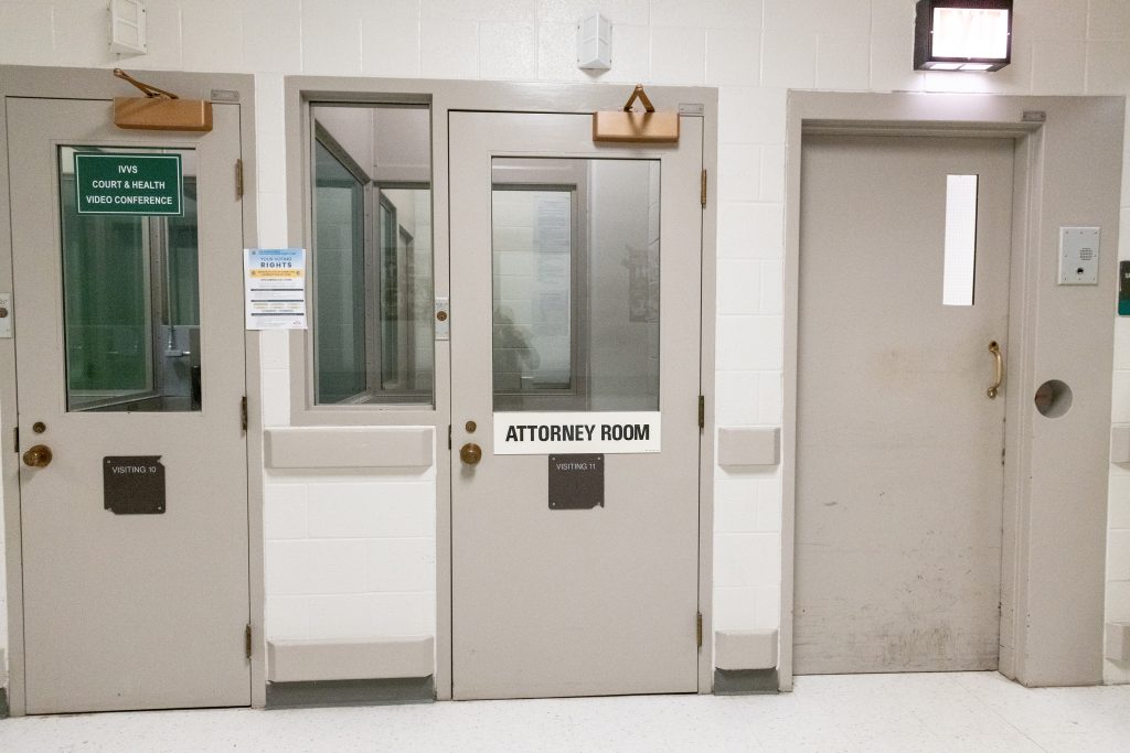 Hallway with gray secured doors, including an attorney room labeled sign; windows with glass panels, posted notices, and overhead lighting on white tile walls
