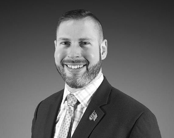 Individual wearing a dark suit jacket and patterned tie with a checkered dress shirt, featuring a lapel pin, posed against a plain gradient background