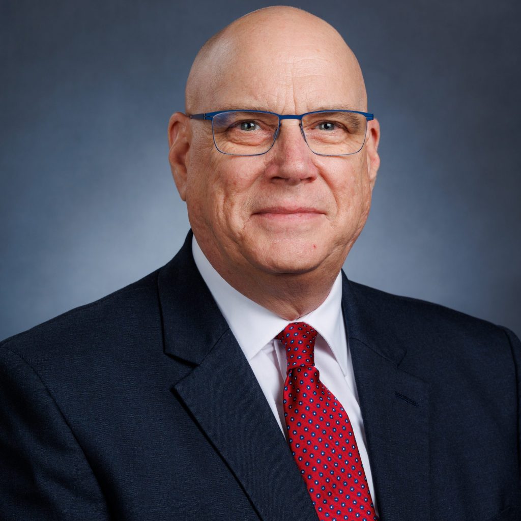 Individual in dark suit jacket, white shirt, and red patterned tie stands before dark blue gradient background.