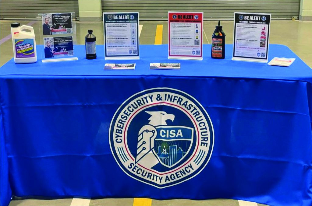 CISA outreach display table covered with blue Cybersecurity and Infrastructure Security Agency cloth, featuring ‘Be Alert’ awareness posters, chemical containers, brochures, QR codes, and public-safety informational materials