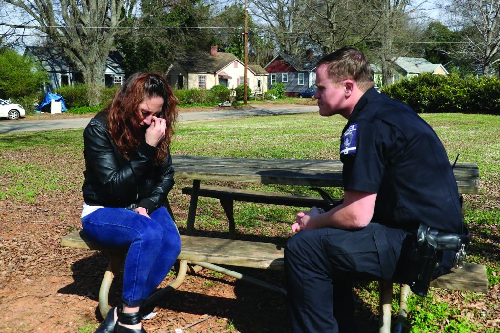 Officer in uniform sits on picnic bench speaking with person in black jacket and jeans; park with trees and houses behind.