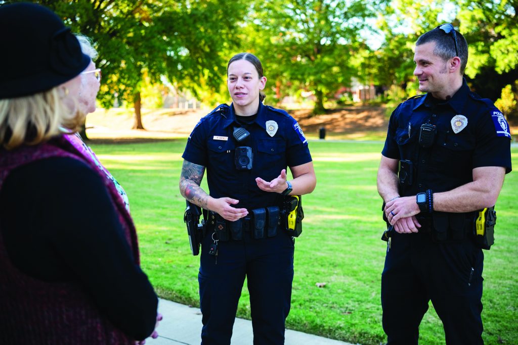 Three uniformed officers stand on grass speaking with a civilian; trees and sunlight in background; all appear engaged in outdoor discussion.