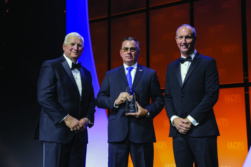 Three individuals in formal black tuxedos and suits stand on stage with orange IACP backdrop; center person holds a clear glass award with blue ribbon accent.