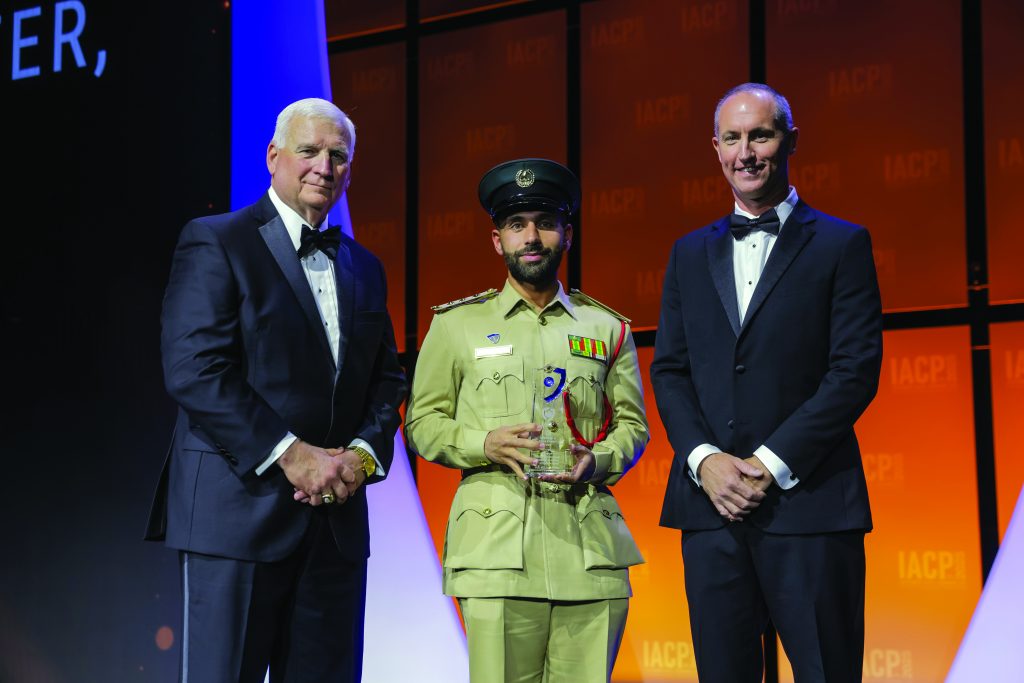 Three individuals stand on stage with orange IACP backdrop; center person in tan military-style uniform with peaked cap holds a clear glass award with blue accent.