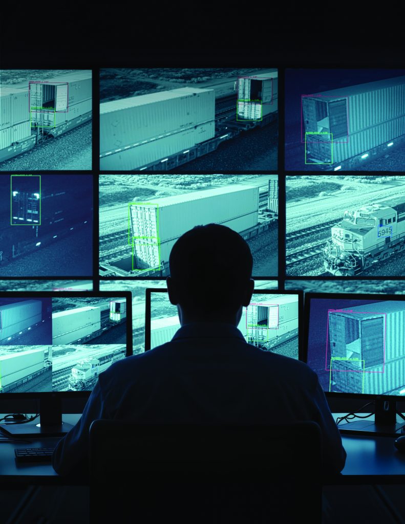 Individual monitors multiple screens showing surveillance footage of cargo containers and train on tracks; screens display various angles and details