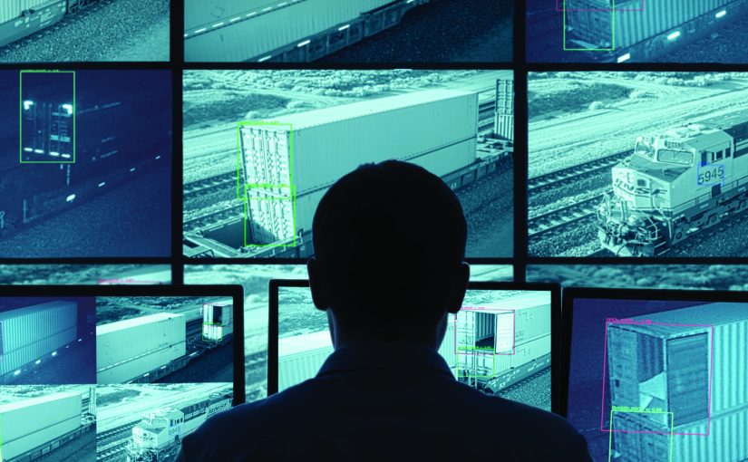 Individual monitors multiple screens showing surveillance footage of cargo containers and train on tracks; screens display various angles and details