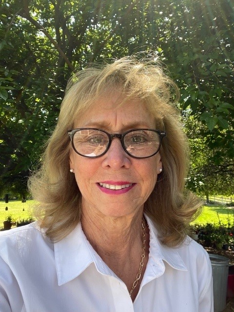 Outdoor portrait of an individual wearing a white collared blouse, glasses, and chain necklace, standing in a sunlit garden with trees, grass, and soft shadows visible