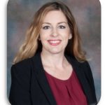 a professional portrait of an individual with long, wavy hair wearing a black blazer over a maroon blouse. The background is a gradient of muted colors, primarily consisting of shades of brown and gray. The photo has rounded corners and appears to be taken in a studio setting with soft lighting that highlights the subject's attire and hair.