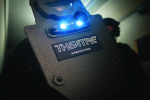 Close-up of tactical ballistic shield labeled THEATRE with bright dual lights illuminated, held by officer in dim hallway during training scenario