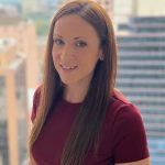 Person wearing a short-sleeve maroon ribbed top with straight, long brown hair, standing on a balcony with blurred city buildings in the background
