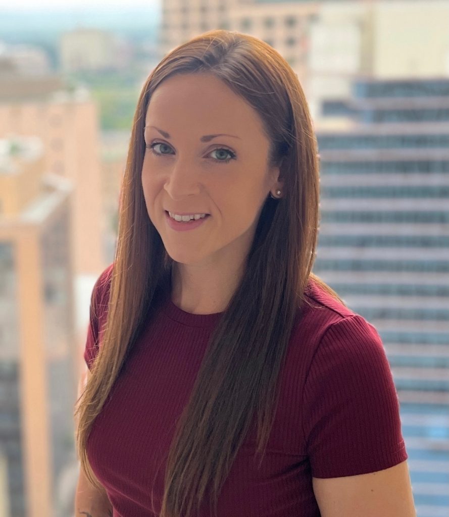 Person wearing a short-sleeve maroon ribbed top with straight, long brown hair, standing on a balcony with blurred city buildings in the background