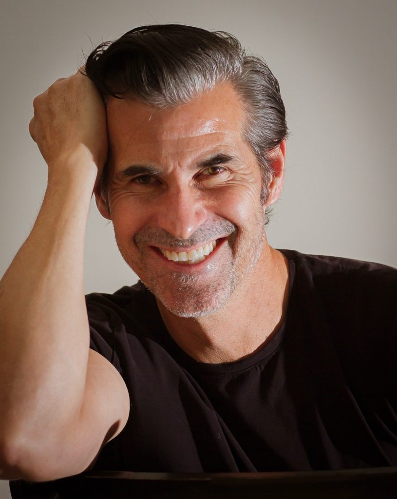 An individual in a dark short‑sleeve shirt sits with one arm bent upward, hand resting on the head, against a softly lit neutral background in a seated portrait pose
