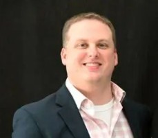 Studio portrait of an individual wearing a dark blazer over a light patterned dress shirt, shown from the chest up against a solid black background with even lighting