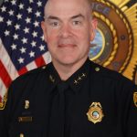 Police official in dark dress uniform with gold badge, collar stars, nameplate and shoulder patches, standing before U.S. flag and large police emblem.
