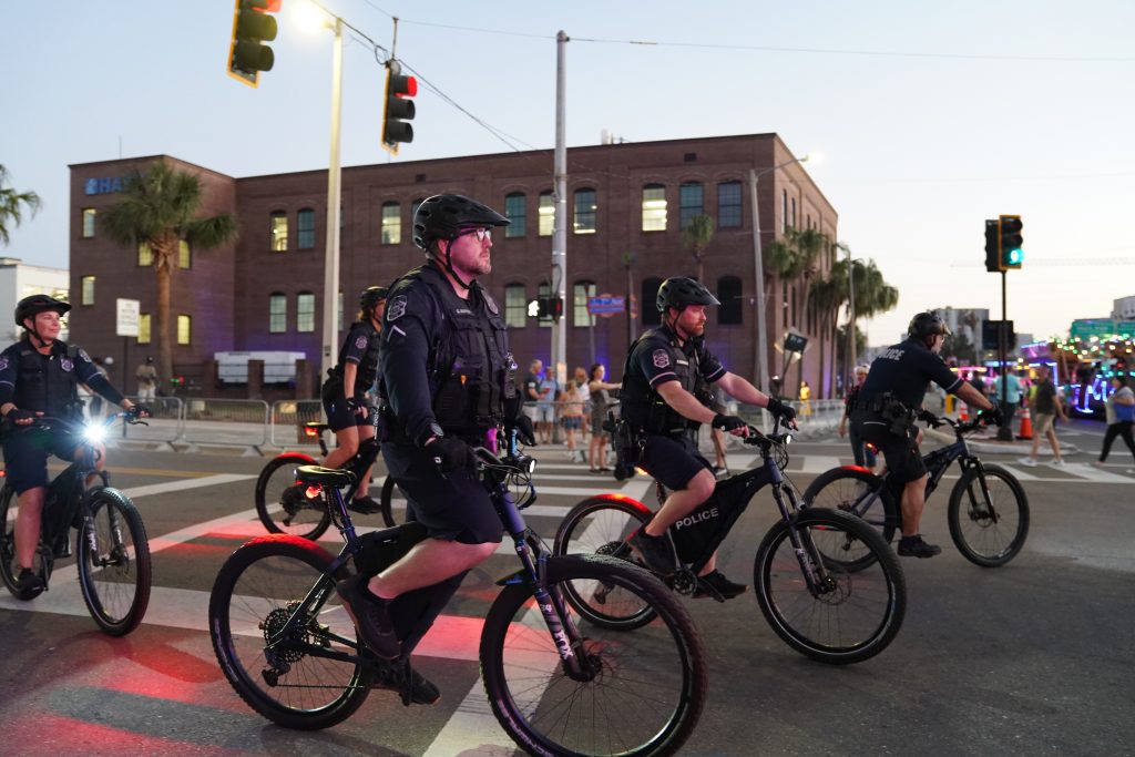 Police bike patrol rides through a busy intersection at dusk, wearing helmets and tactical gear, with brick buildings, traffic lights, and event crowds in the background.