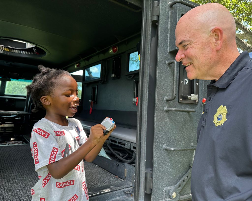 Child holding small toy stands inside armored police vehicle talking with uniformed officer at open door, with interior benches and equipment visible.