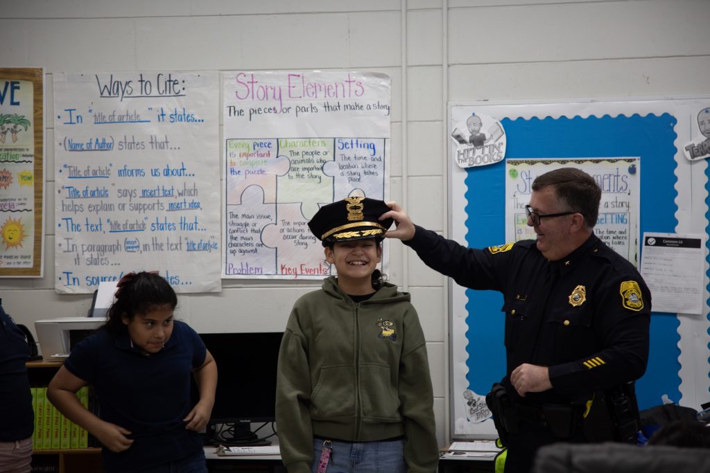 Police officer in dress uniform places a police hat on a student in a classroom, with posters on writing skills and story elements displayed on the wall behind them.