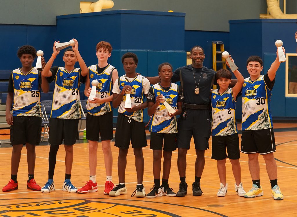 Youth basketball team in blue and yellow Storm jerseys stands in a gym holding trophies beside a uniformed officer, bleachers visible behind.