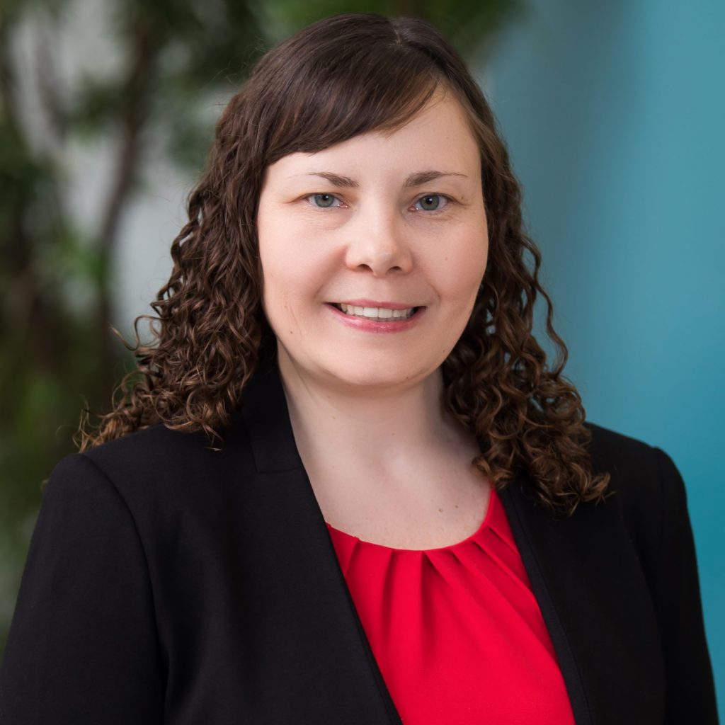 a woman with curly brown hair wearing a black blazer over a red blouse. The background is blurred, showing some greenery and a light blue wall.