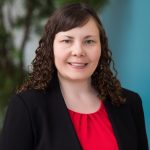 a woman with curly brown hair wearing a black blazer over a red blouse. The background is blurred, showing some greenery and a light blue wall.
