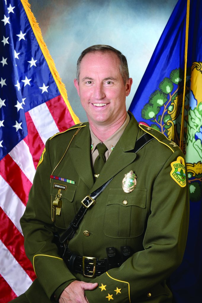 Law enforcement officer in formal green uniform with badges, shoulder patch, and service ribbons, seated between U.S. and state flags on studio backdrop