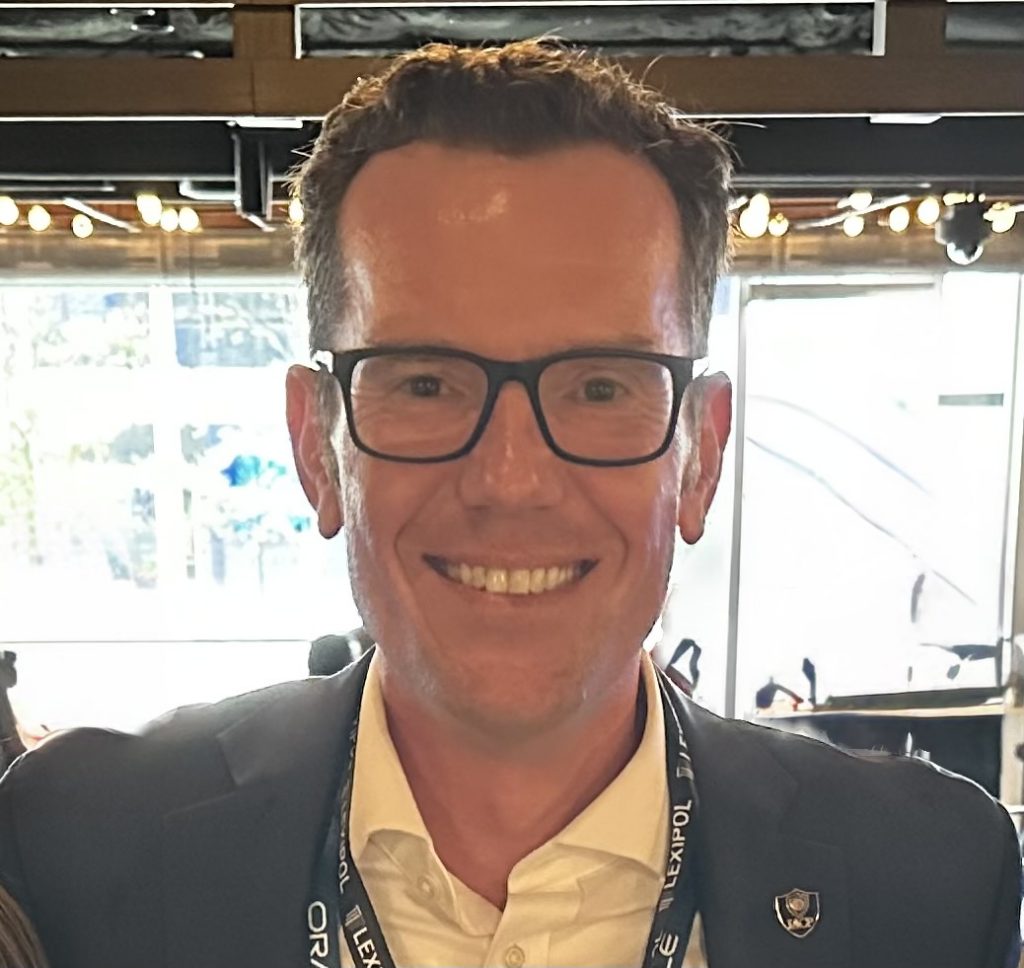 Individual in white shirt and dark blazer with badge on lapel and 'LEXIPOL' lanyard stands indoors; background blurred