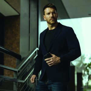 Person in black blazer and jeans holds smartphone near metal railing; urban background with buildings, trees, and soft natural lighting.