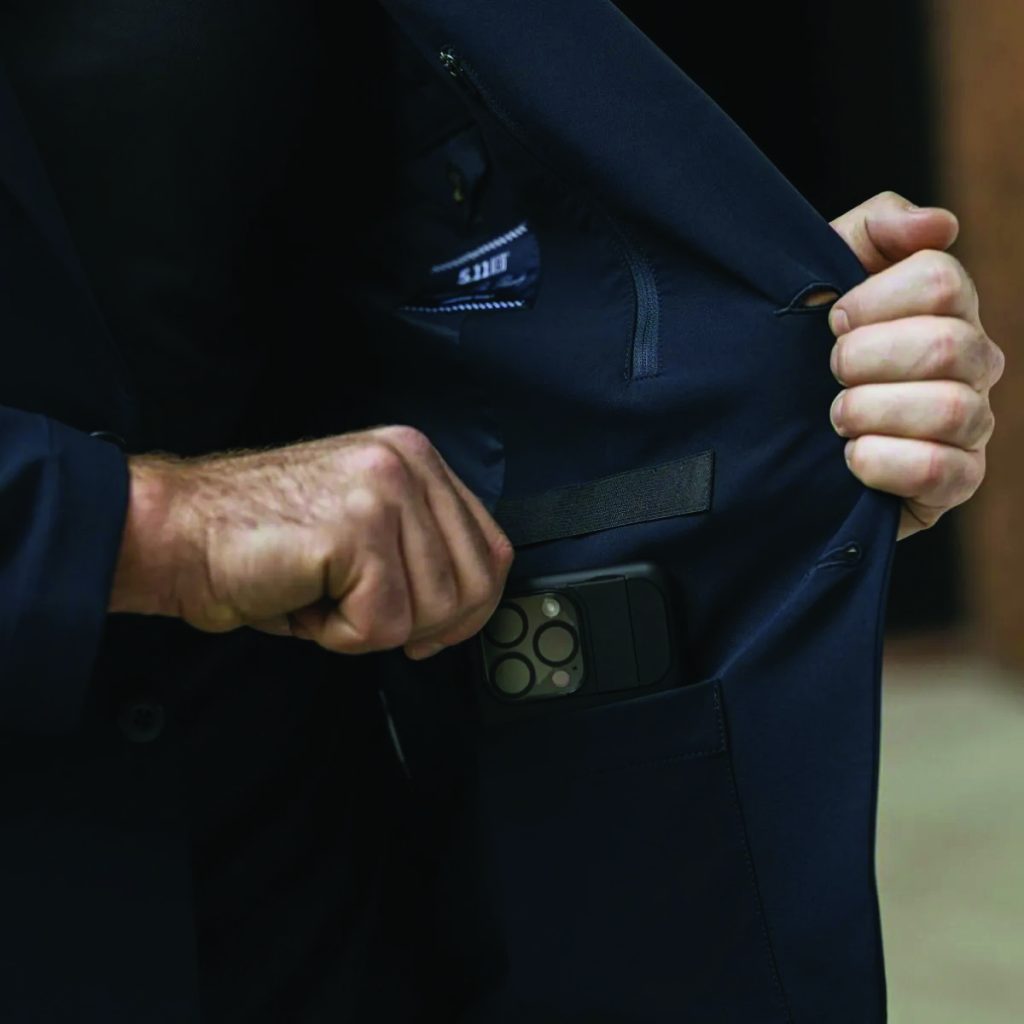Person in dark suit jacket holds it open to reveal an iPhone with quad-camera in inner pocket; background is blurred and indistinct.