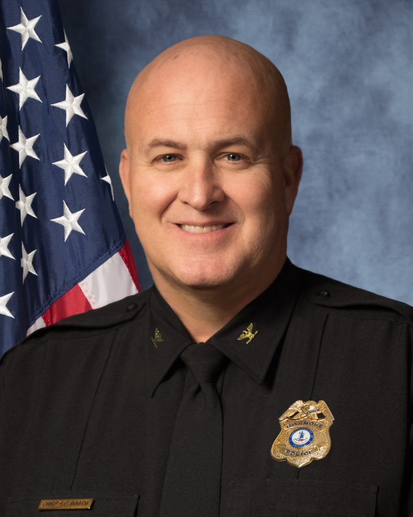 Uniformed officer in black dress shirt with gold badge and nameplate, standing before an American flag and gray backdrop