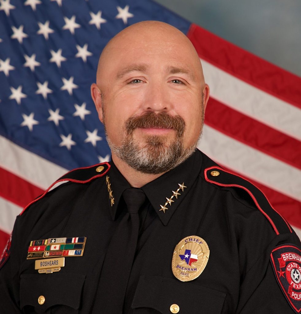 Police chief in black uniform with gold stars on collar, service ribbons, nameplate, badge, and red‑trimmed shoulder patch stands before a large American flag backdrop.