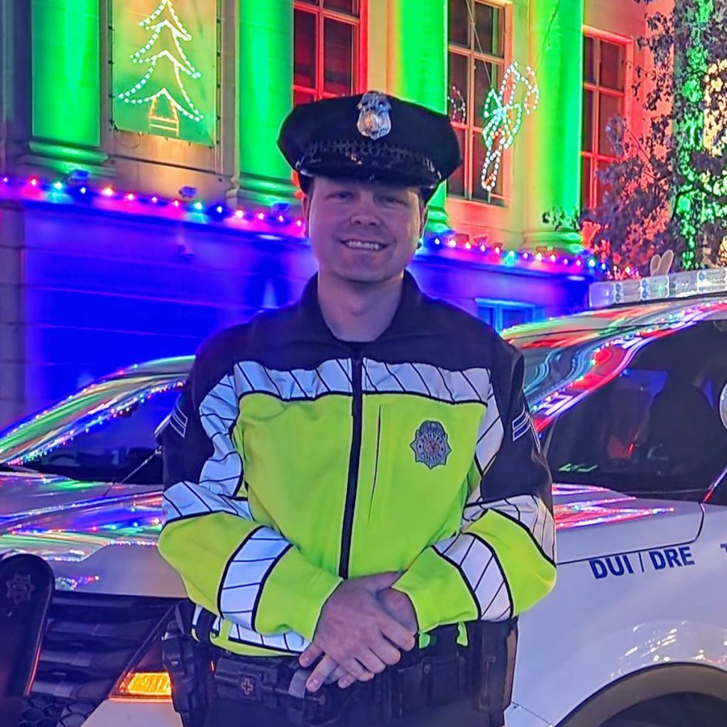 Officer in high-vis jacket stands before two 'DUI/DRE Traffic' patrol cars parked outside brightly lit building adorned with holiday string lights