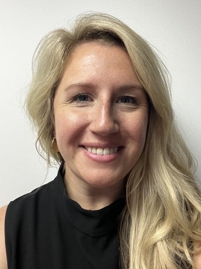 Studio portrait of an individual wearing a sleeveless black top and small hoop earrings, shown from shoulders up against a plain light wall with soft, even lighting