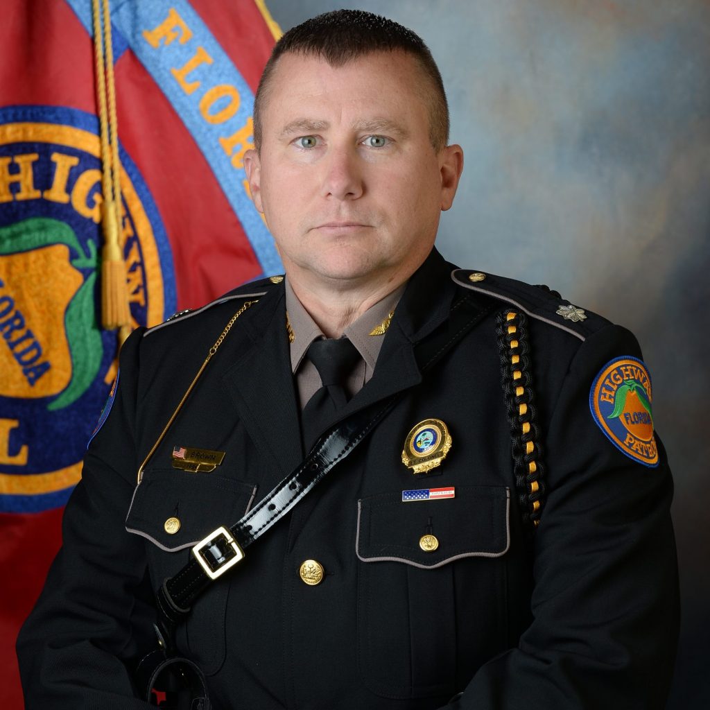 Uniformed officer with badges stands before red flag bearing Florida Highway Patrol emblem; focus on insignia, uniform details, and official setting