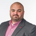 Person in grey blazer with red pin, red checkered shirt, and white undershirt stands against plain white background; face not visible.