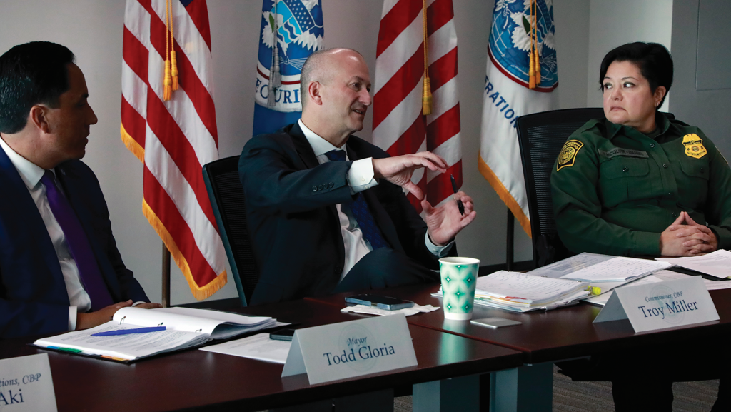 Three individuals seated at conference table with nameplates 'Todd Gloria' and 'Troy Miller'; U.S. flags and Homeland Security banners in background