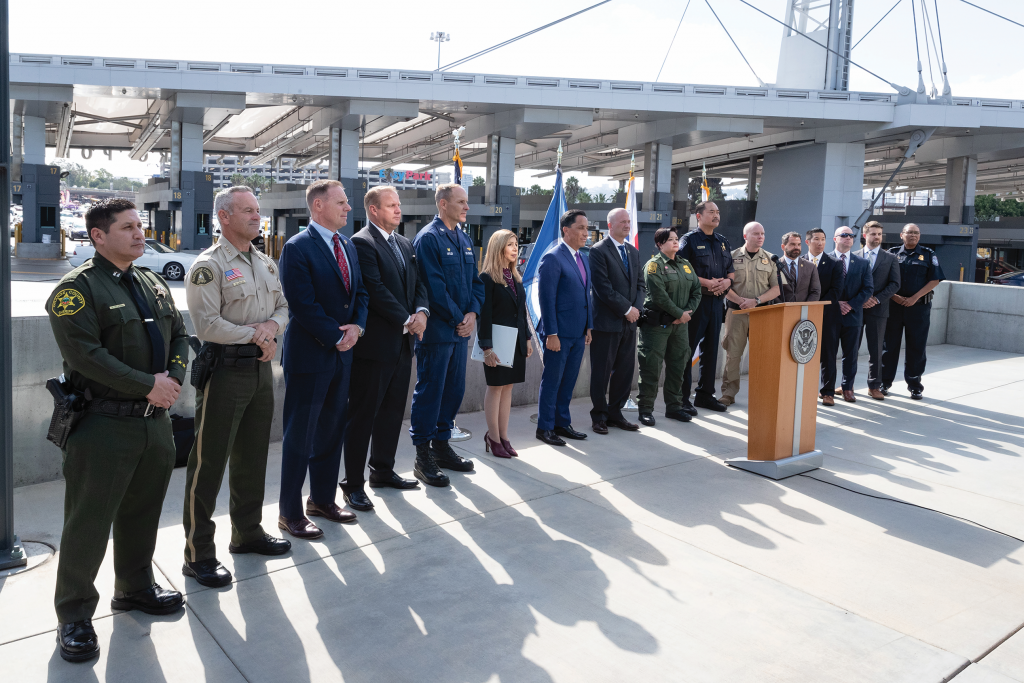 Group of law enforcement officials in uniforms and suits stand beside podium with emblem; modern bridge structure with pillars and cables in background