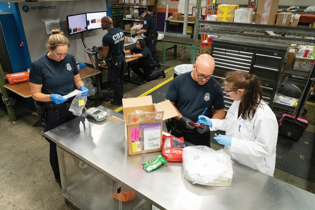 Law enforcement and lab technician inspect boxes and bags on metal table in warehouse; various packages and materials under examination