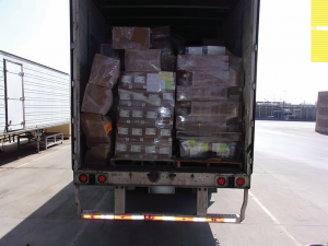Open truck trailer at loading dock filled with assorted boxes and plastic-wrapped pallets; adjacent trailer visible—potential inspection or transit point