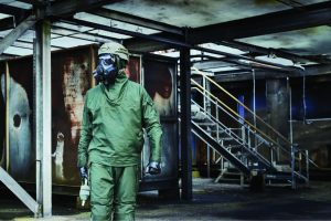 Person in full green CBRN protective suit with respirator and helmet walks through industrial structure with steel beams, stairs, rusted tanks
