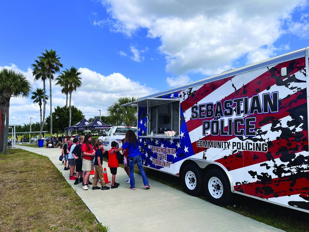 a group of children standing in line outside a large trailer with "Sebastian Police Community Policing" written on the side. The trailer has a patriotic design with red, white, and blue colors and stars. Some children are wearing red shirts, while others are in blue. An adult in a blue shirt is interacting with the children near the entrance of the trailer. In the background, there are palm trees and another tent or booth set up with people around it. The sky is partly cloudy with patches of blue visible. The faces of the children and the adult are visible, showing various expressions of interest and engagement.