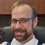 Person seated in an office wearing a white dress shirt, blue patterned tie, and blue suspenders, with wooden cabinets and books visible in the background
