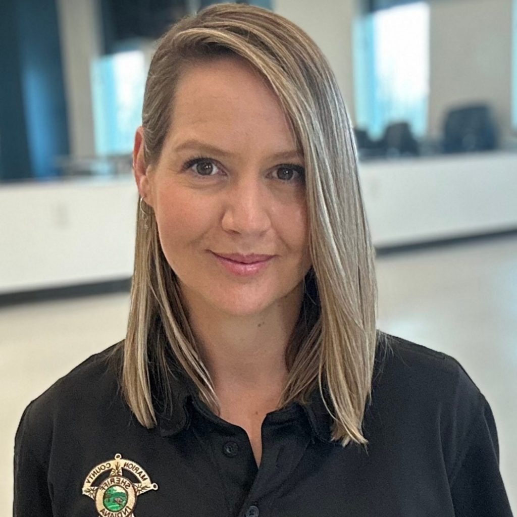 Individual in black polo with sheriff’s badge on left chest stands indoors; background features large windows and softly blurred architectural elements