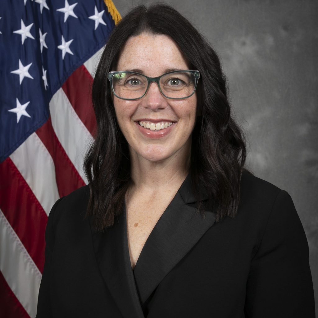 Person with shoulder-length dark hair in black suit and shirt stands before U.S. flag; gray textured wall forms the background.